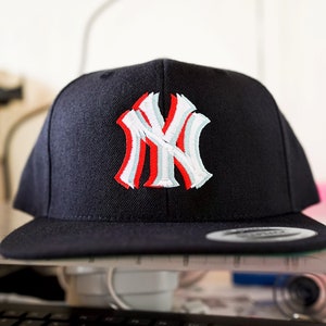 Puede incluir: Una gorra de béisbol negra con un logotipo bordado de los New York Yankees en blanco y rojo.