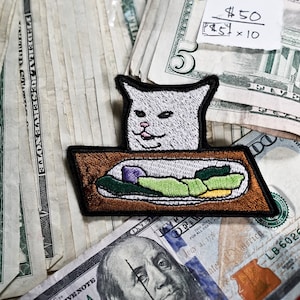 Op de afbeelding: Een witte kat meme-patch met een zelfvoldane uitdrukking, zittend op een bruin dienblad met groene en gele groenten. De patch ligt op een stapel Amerikaanse dollarbiljetten, waaronder een biljet van $100.