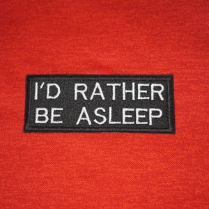Peut inclure: Sweat-shirt rouge avec un patch noir qui dit "I'd rather be asleep" en texte blanc.