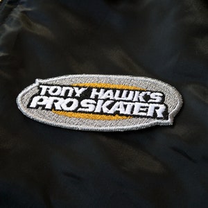 Könnte beinhalten: Schwarze Jacke mit goldenem Reißverschluss und einem Aufnäher mit der Aufschrift "TONY HAWK'S PRO SKATER" in weißen und gelben Buchstaben. Der Aufnäher ist grau umrandet und hat einen schwarzen Hintergrund.