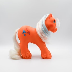May include: Orange toy pony with white and blue streaked mane and tail. The pony has blue eyes and a design on its flank with a blue triangle, red bow, and crescent moon. A vintage collectible toy.