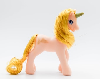 May include: A vintage pink unicorn toy with yellow hair and tail. The unicorn has a green horn and blue eyes. The toy is standing on all four legs, with a light-colored background.