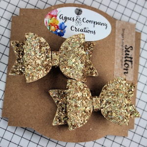 May include: Two gold glitter hair bows on a brown card with the text "Agnes & Company Creations" and "Sutton". The bows are in a classic bow shape, perfect for adding sparkle to any hairstyle.