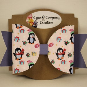 May include: A handmade hair bow featuring a festive penguin pattern on a light pink background. The bow has purple accents and is mounted on a brown card with the text "Agnes & Company Creations". The penguins wear hats and scarves.