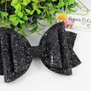 May include: A large, black glitter bow with a layered design. The bow is centered in the image, with a bright green leafy background and a small business card that reads "Agnes & Company Creations".