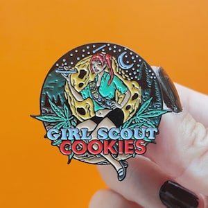 May include: A round enamel pin featuring a Girl Scout holding a tray of cookies in front of a moon and forest scene. The pin has the text "GIRL SCOUT COOKIES" in a stylized font, with cannabis leaves around the moon.