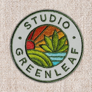 May include: Embroidered patch with a circular design featuring the text "STUDIO GREENLEAF". The patch has a light gray background with a green border. The center design includes a stylized landscape with a red sun, green plants, and blue water.