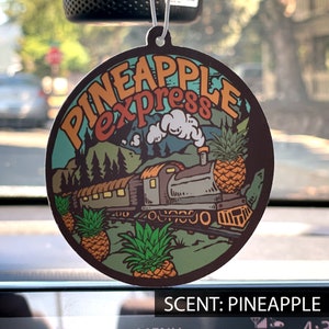 May include: Round air freshener with a train and pineapple graphic. The text "PINEAPPLE express" is in orange and brown. The scent is pineapple.