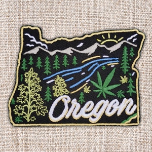 Peut inclure: Écusson brodé en forme de l'Oregon, avec un fond noir et une bordure jaune. Le motif comprend des montagnes, des arbres, une rivière, une feuille de cannabis et le mot "Oregon" en écriture blanche.