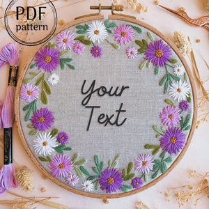 以下が含まれることがあります： 灰色のリネン生地の背景を持つ木製刺繍枠。生地には、紫と白の花のリースが刺繍されています。枠の中央には「Your Text」という文字が刺繍されています。