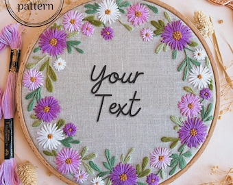 May include: A wooden embroidery hoop with a gray linen fabric background. The fabric is embroidered with a wreath of purple and white flowers. The text "Your Text" is embroidered in the center of the hoop.