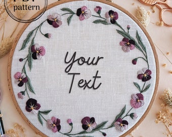 May include: A white embroidery hoop with a floral design of purple and green pansies. The text "Your Text" is embroidered in the center of the hoop.