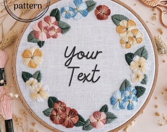 May include: A white embroidery hoop with a floral wreath design. The wreath is made up of pink, yellow, blue, and brown flowers with green leaves. The text "Your Text" is in the center of the hoop.