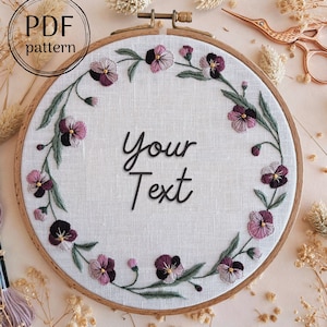 May include: A white embroidery hoop with a floral design of purple and green pansies. The text "Your Text" is embroidered in the center of the hoop.