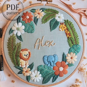 Könnte beinhalten: Gesticktes Stickrahmen-Kunstwerk mit Dschungel-Thema, einschließlich Löwe, Giraffe und Elefant. Der Name "Alex" ist in der Mitte gestickt. Das Design umfasst Blumen und Blätter in kreisförmiger Anordnung. Der Text "PDF pattern" befindet sich oben links.