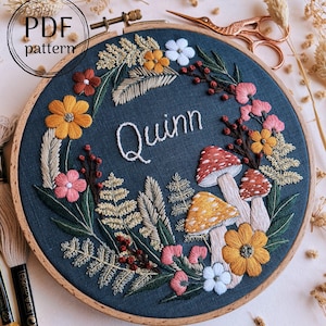 May include: Embroidered hoop art with a dark blue background featuring a floral wreath with mushrooms, leaves, and the name "Quinn" in white thread.