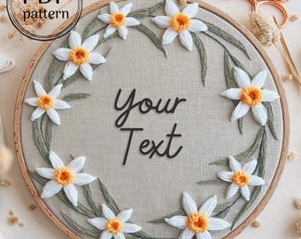 May include: A white embroidery hoop with a beige linen fabric background. The fabric is embroidered with a wreath of white daffodils with yellow centers and green leaves. The text "Your Text" is embroidered in black in the center of the hoop.