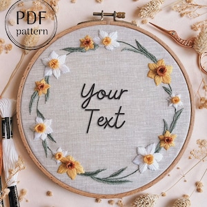 May include: A white embroidery hoop with a beige linen fabric background. The fabric is embroidered with a wreath of yellow and white daffodils. The text "Your Text" is embroidered in the center of the hoop.
