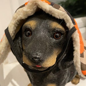 Peut inclure: Un chien noir et brun portant un manteau de chien à capuche camouflage et orange avec une doublure en polaire blanche.