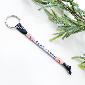 May include: Black and white keychain with pink and gold beads spelling "HOCKEY MOM" with a heart symbol between the words.