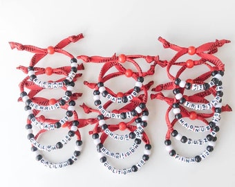 May include: A set of 12 red and black hockey bracelet style friendship bracelets with white letters spelling out names and numbers. The bracelets are made of red and black string with black beads and white letter beads.