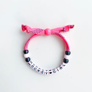 Peut inclure: Bracelet en perles rose et noires avec les mots "HOCKEY GIRL" et un charme en forme de cœur.