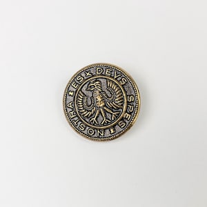 Könnte beinhalten: Eine runde Metallmünze im antiken Stil mit einem detaillierten Emblem eines Vogels und der Inschrift "SPES DEIS NOSTRA". Die Münze hat eine Bronze- und Silberfarbgebung mit aufwendigen Details an den Rändern und dem zentralen Design.