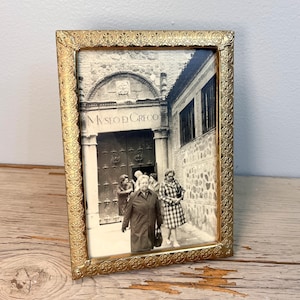 May include: A vintage gold-colored picture frame with an ornate, embossed border. The frame holds a black and white photograph of a building with the text "MUSEO D GRECO" above the entrance. The photograph features several people.