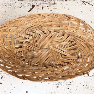 May include: Oval-shaped woven basket in natural tan color. The basket is made of interwoven strips of wood, creating a textured surface. The basket's design features a central starburst pattern.