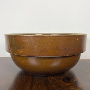 May include: A brown ceramic bowl with a wide, flat rim and a rounded base. The bowl has a two-tiered design, with the rim forming a distinct upper level. The surface has a slightly glossy finish, and the color is a warm, earthy brown.