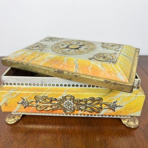 May include: An antique jewelry box with a hinged lid, featuring a yellow and white marble design. The box has gold-colored metal trim and decorative accents, including ornate feet. The lid is slightly ajar, revealing the interior.