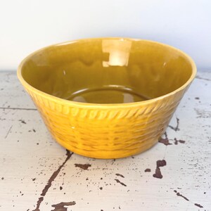 May include: A mustard yellow ceramic bowl featuring a textured exterior that mimics the appearance of a woven basket. The bowl has a smooth, glossy interior finish and a distinct rope-like pattern encircling the upper rim of the vessel.