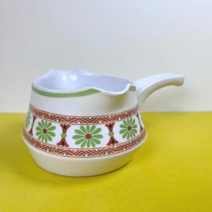 May include: Cream ceramic creamer with a handle and a decorative floral pattern in green, red, and brown. The creamer has a green band around the top rim. The creamer is on a yellow surface.