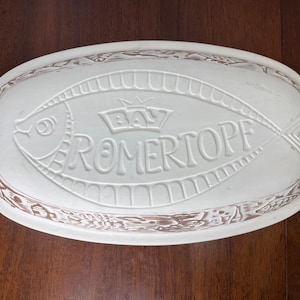 May include: Off-white oval baking dish with a fish design and the text "BAY ROMERTOPF". The dish has handles on each end and a decorative brown border. A crown is above the text.