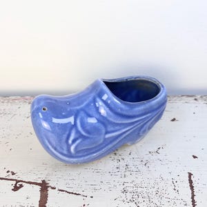 May include: A periwinkle blue ceramic clog-shaped planter or decorative vessel. The object features a glossy finish and a molded relief design of a tulip on the side. It has a hollow interior and a small circular hole near the toe area.