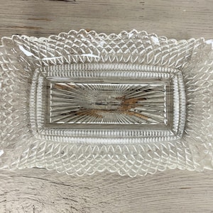 May include: A clear, rectangular glass dish with a decorative, diamond-patterned border. The center features a sunburst design. The dish is likely used for serving or display.