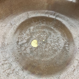 May include: Close-up of a textured, bronze-colored bowl with a circular indentation in the center. A small, gold-colored spot is visible in the center of the bowl. The bowl has a worn, aged appearance.