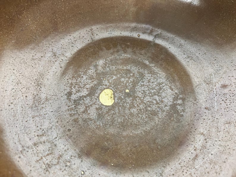 May include: Close-up of a textured, bronze-colored bowl with a circular indentation in the center. A small, gold-colored spot is visible in the center of the bowl. The bowl has a worn, aged appearance.