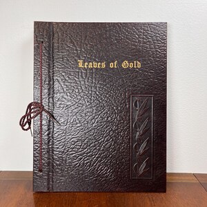 以下が含まれることがあります： 「Leaves of Gold」の金色の文字が入った、濃い茶色の質感のあるフォトアルバム。右側には葉のデザインの装飾的な長方形のパネルがあります。茶色の紐でアルバムが閉じられています。