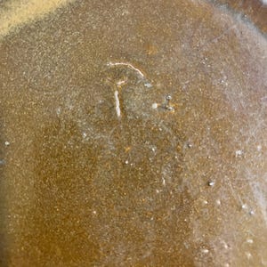 May include: Close-up of a brown ceramic plate with a textured surface. The plate has a light-colored rim and a small, white, stylized letter "T" etched into the center. The plate's surface has small, white specks.