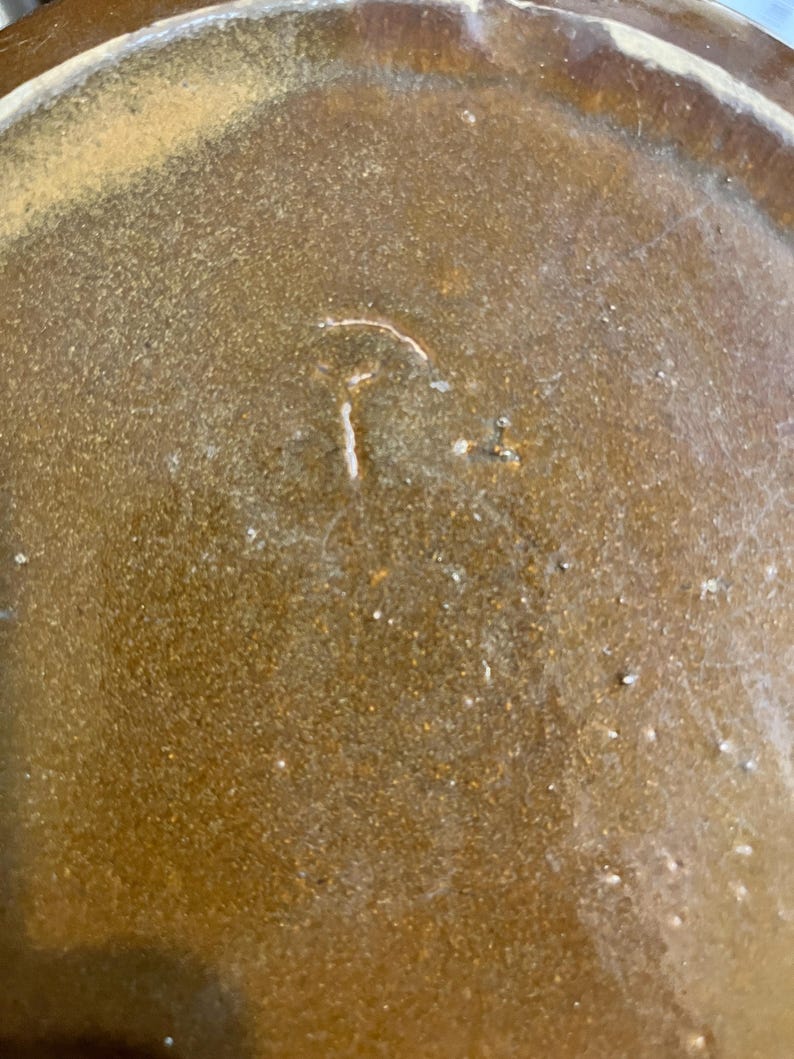 May include: Close-up of a brown ceramic plate with a textured surface. The plate has a light-colored rim and a small, white, stylized letter "T" etched into the center. The plate's surface has small, white specks.