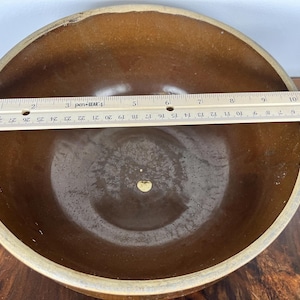 May include: A brown ceramic bowl with a light tan rim. A ruler is placed across the bowl, measuring approximately 10 inches in diameter. The bowl has a smooth, glossy interior.
