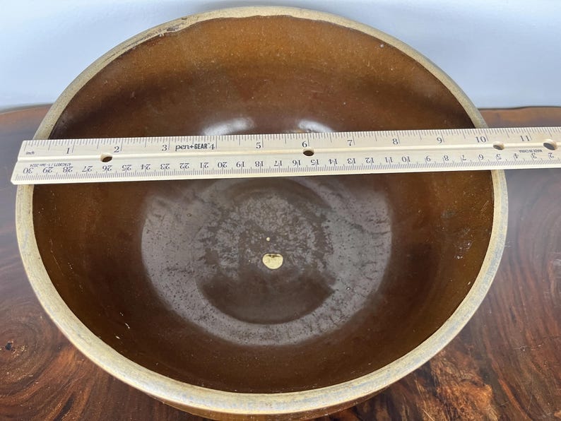 May include: A brown ceramic bowl with a light tan rim. A ruler is placed across the bowl, measuring approximately 10 inches in diameter. The bowl has a smooth, glossy interior.