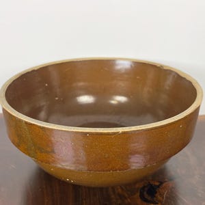 May include: A brown ceramic bowl with a rounded shape. The bowl has a wide opening and a slightly textured surface. The rim of the bowl is a lighter shade of brown. The bowl is sitting on a wooden surface.
