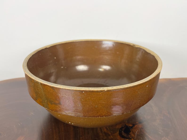 May include: A brown ceramic bowl with a rounded shape. The bowl has a wide opening and a slightly textured surface. The rim of the bowl is a lighter shade of brown. The bowl is sitting on a wooden surface.