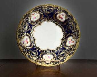 May include: A decorative plate with a navy blue and gold design. The plate features a white center and is adorned with floral accents. The rim is gold, and the plate is displayed on a gold stand. The plate has a detailed, ornate pattern.