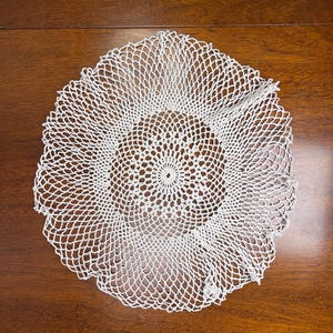May include: A white, circular doily with a delicate, openwork crochet design. The doily features a central, tightly crocheted circle that expands into a lacy, scalloped edge. The intricate pattern is visible against a dark wooden surface.