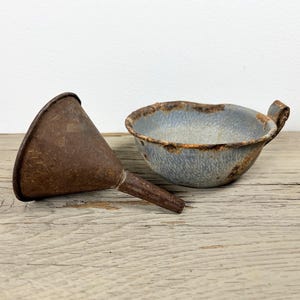 May include: An antique metal funnel and a small bowl with a handle. The funnel is brown and rusty, with a conical shape. The bowl is blue with a speckled pattern and a rusty rim. Both items are placed on a wooden surface.