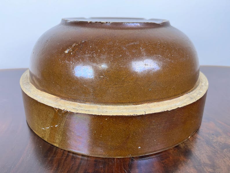 May include: A brown ceramic crock with a rounded top and a wide, cylindrical base. The crock has a glossy finish and a light-colored band around the base. The antique kitchenware is likely used for food storage.