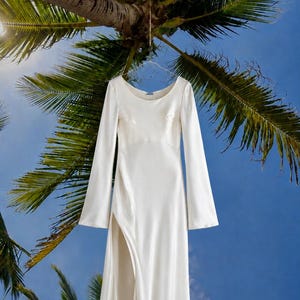 以下が含まれることがあります： 長袖の白いドレス。ラウンドネック、体にフィットするボディス、そしてサイドに深いスリットが入ったロングスカートが特徴です。ハンガーに吊るされたこのドレスは、全体にほのかな光沢がある滑らかな無地の生地で作られています。